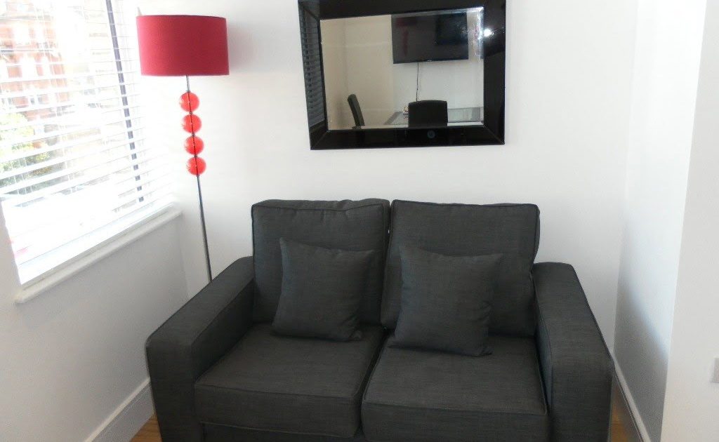 A modern room featuring a dark gray two-seater couch, a black-framed mirror, a red floor lamp, red decorative balls, and a white blind-covered window.