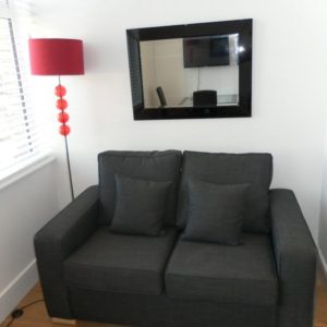 A modern room featuring a dark gray two-seater couch, a black-framed mirror, a red floor lamp, red decorative balls, and a white blind-covered window.