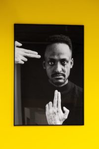 A framed black and white photograph of a person with intense gaze, framed by two hands, against a bright yellow background.