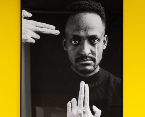 A framed black and white photograph of a person with intense gaze, framed by two hands, against a bright yellow background.