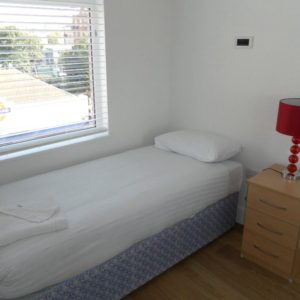 A minimalistic bedroom with a single bed, white bedding, a red lamp on a wooden nightstand, and a window with horizontal blinds.