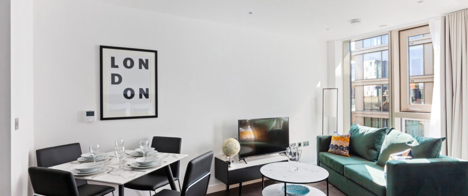 Modern living room with natural light, white walls, green sofa, marble tables, a dining area, and a 'LONDON' framed artwork. Elegant and tidy space.