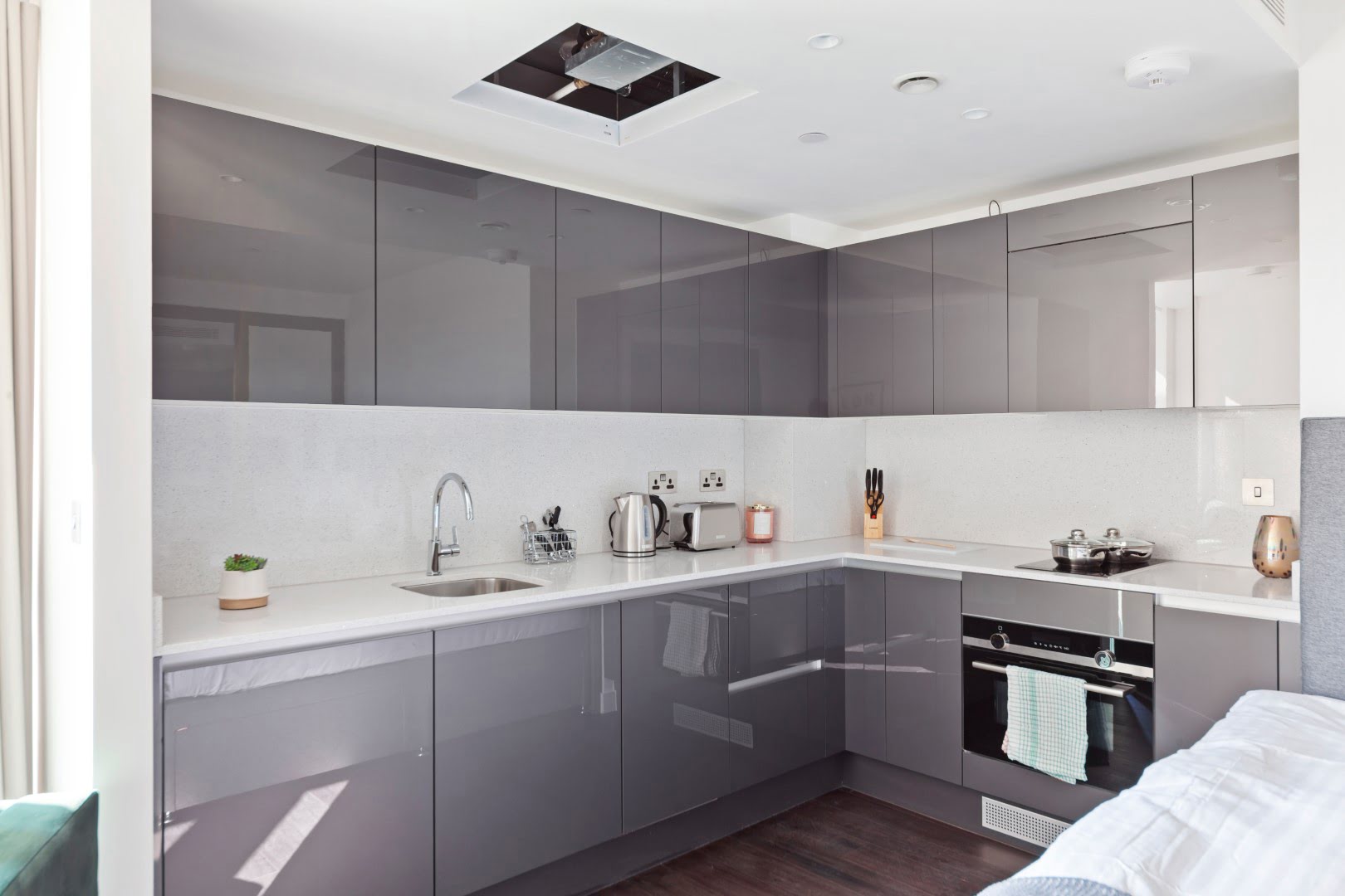 This image shows a modern kitchen with gray cabinets, white countertops, recessed lighting, stainless steel appliances, and a sprinkling of domestic accessories.