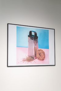 A framed image on a wall displays a cheese grater with shavings beside a round, crumbled object on a pink and blue background.