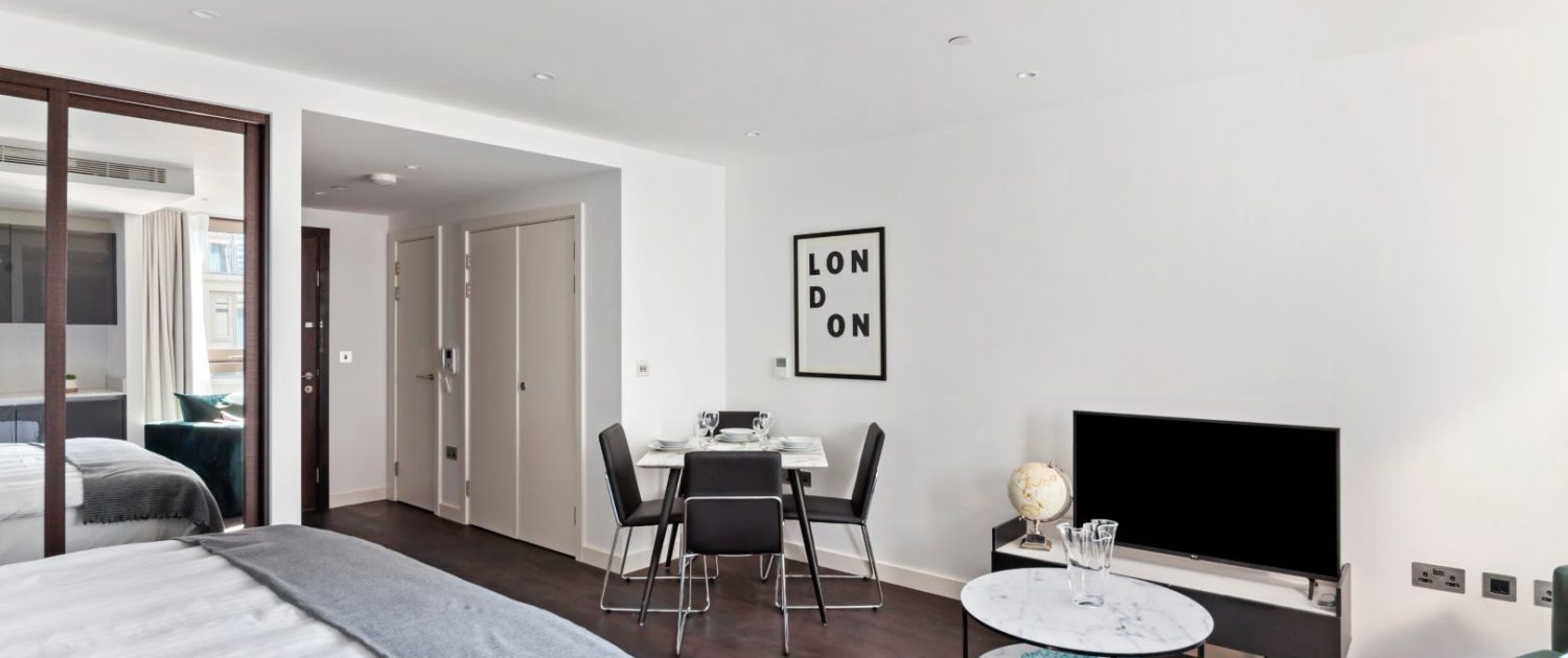 Modern studio apartment with dining area, bed, and living space. Includes a "LONDON" wall art, globe, dark wood flooring, and neutral color palette.