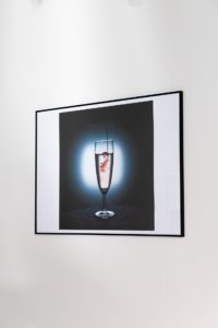 A framed photograph on a white wall depicts a champagne flute with effervescent liquid and splashing droplets, emphasizing motion and contrast with a dark background.