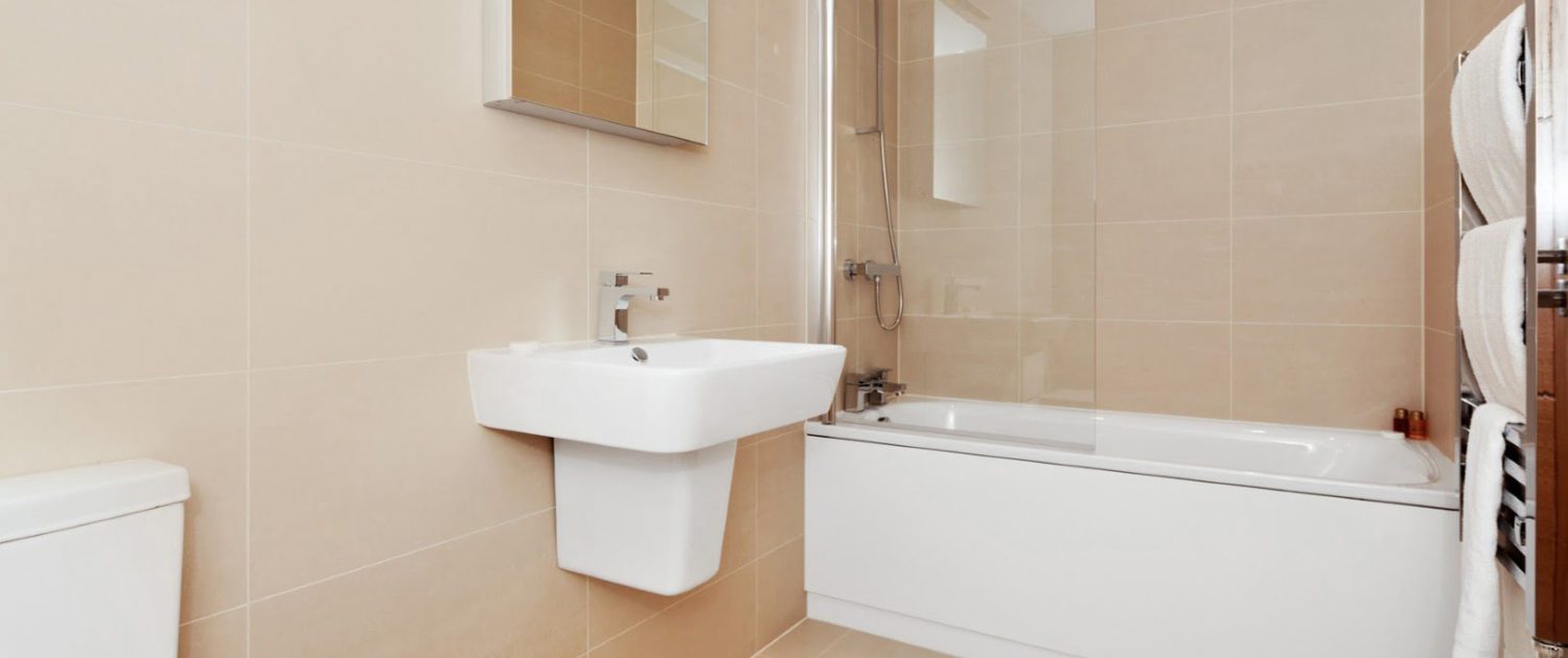 A clean, modern bathroom with beige tiles, featuring a white toilet, sink, bathtub with shower, mirror, and a heated towel rail.