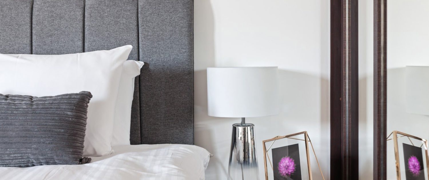 A modern bedroom with a gray upholstered headboard, crisp white bedding, a chic table lamp, and a mirror reflecting a framed flower photograph.