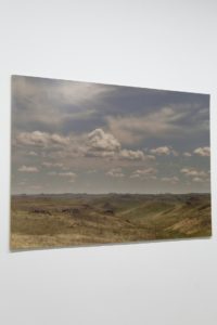 This image showcases a landscape canvas print affixed to a wall. It depicts rolling hills under a vast sky with dynamic clouds. The scene conveys serenity and open space.