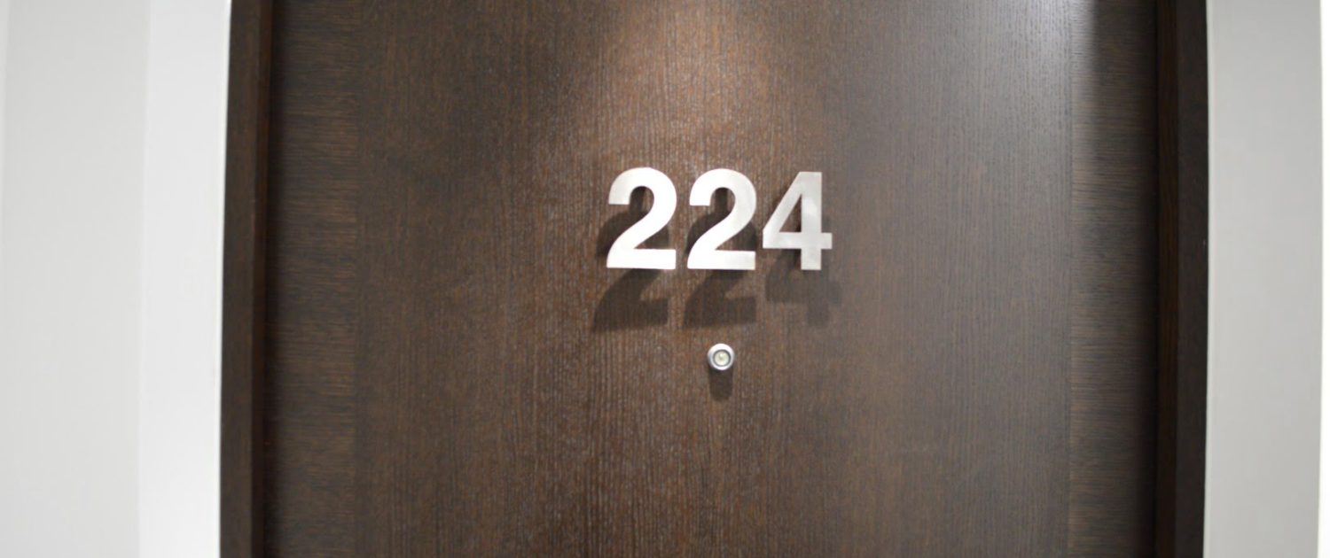 This is a dark wooden door with the number 224 in silver, raised numerals. There's a peephole and a light shadow cast on the surface.