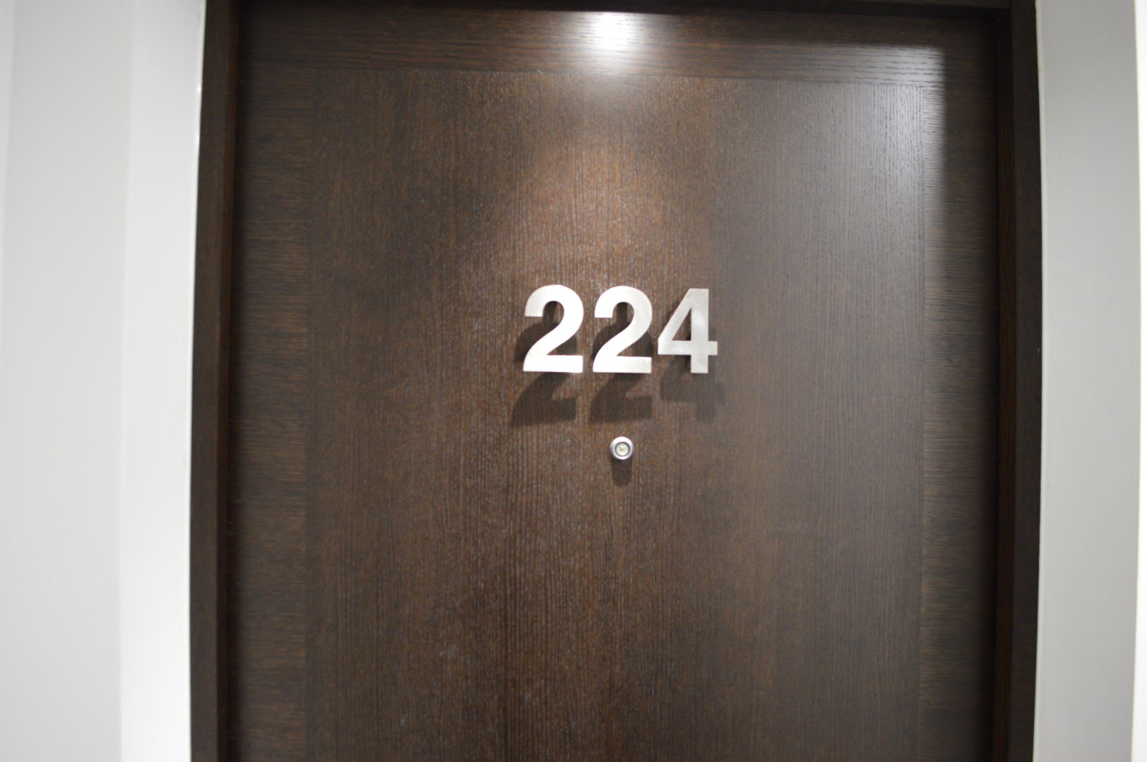 This is a dark wooden door with the number 224 in silver, raised numerals. There's a peephole and a light shadow cast on the surface.