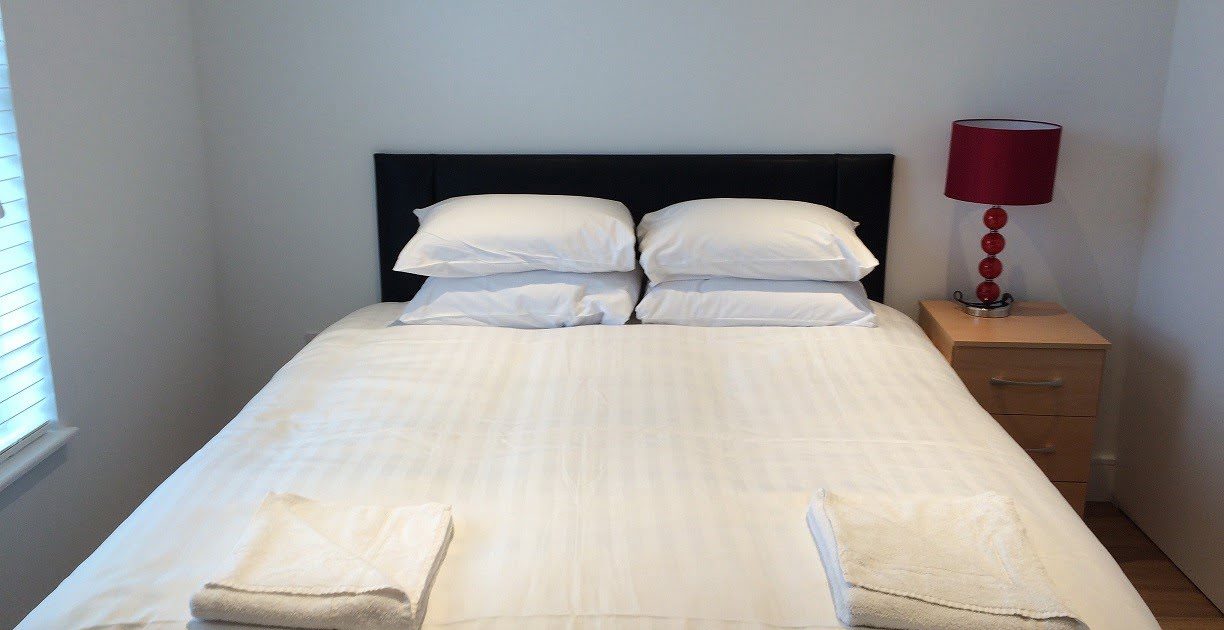 A neatly made bed with white bedding, two pillows, and towels placed at the foot. A nightstand with a red lamp is beside the bed.