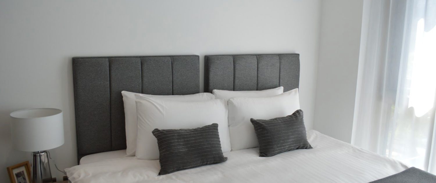 A neatly made bed with white bedding, gray upholstered headboard, two pillows, two gray accent cushions, and a minimalist nightstand with a lamp.