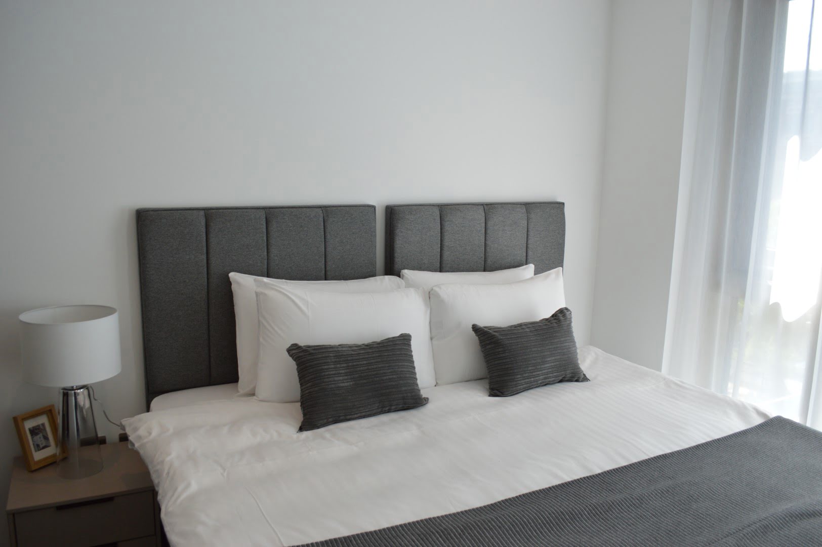 A neatly made bed with white bedding, gray upholstered headboard, two pillows, two gray accent cushions, and a minimalist nightstand with a lamp.