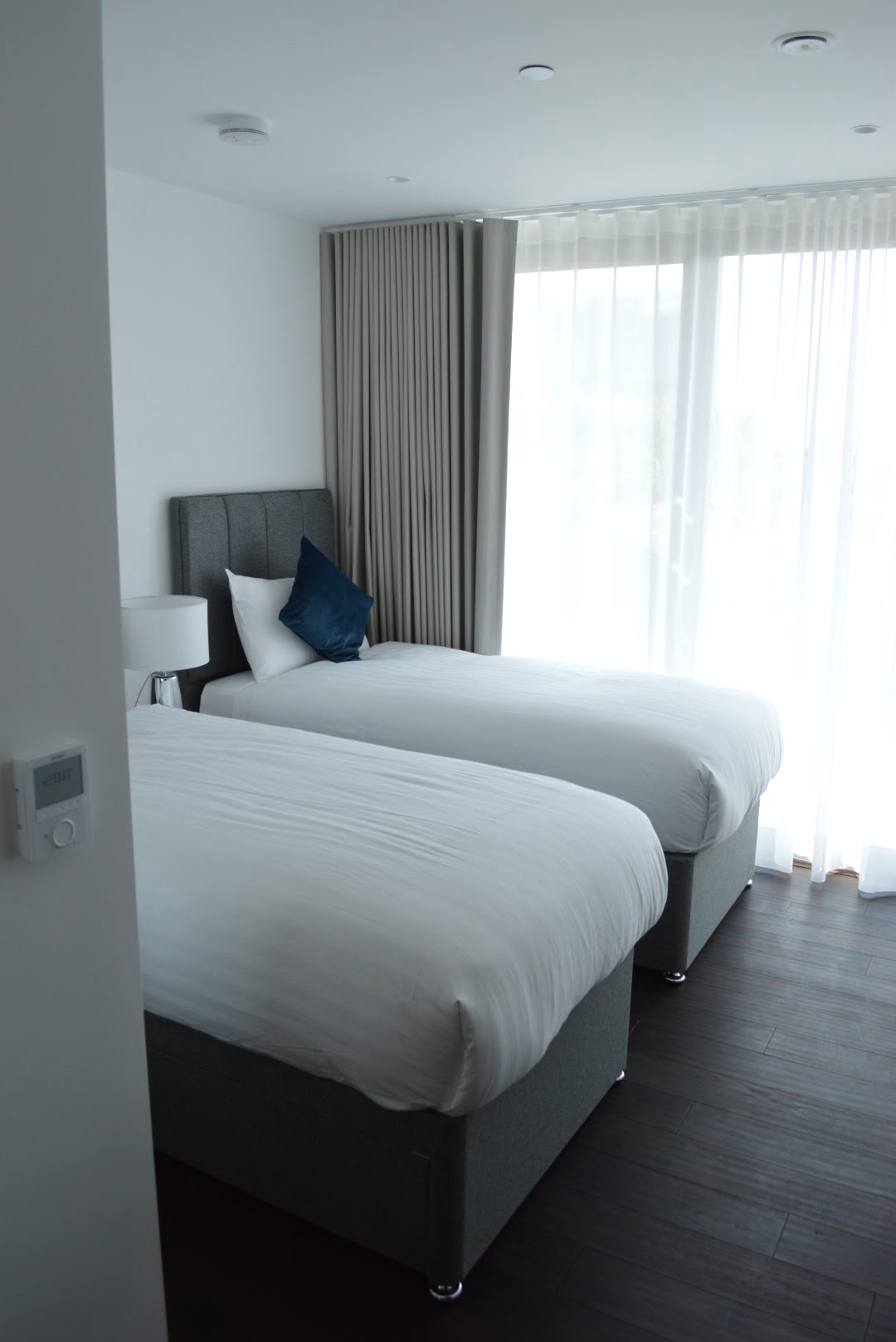 A modern, minimalist hotel room with two single beds, white bedding, a grey headboard, and sheer curtains in front of a window. Dark wood flooring contrasts the light walls.