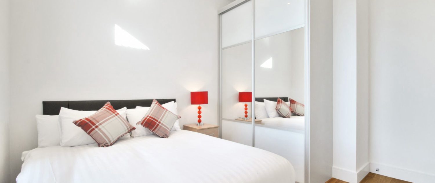 A modern, minimalist bedroom featuring a large bed with white bedding, plaid pillows, red bedside lamps, and a mirrored sliding-door wardrobe.