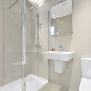 A modern, clean bathroom with a glass shower cubicle, a white pedestal sink, a mirror, a toilet, and beige wall tiles.