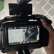 A person's hand is holding a professional video camera with a screen showing a black and white image of a person's face, implied to be the subject being filmed.