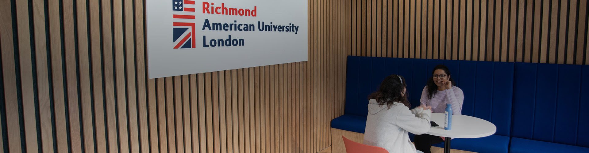 Two people are seated opposite each other at a white table inside a modern room with wooden slats and a sign for Richmond American University London.
