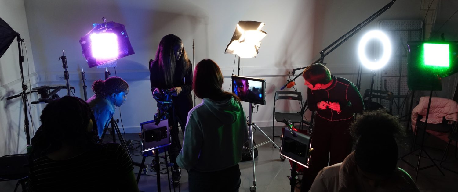 A film or video production scene with various people working around professional lighting equipment, cameras, and a monitor in a dark room.