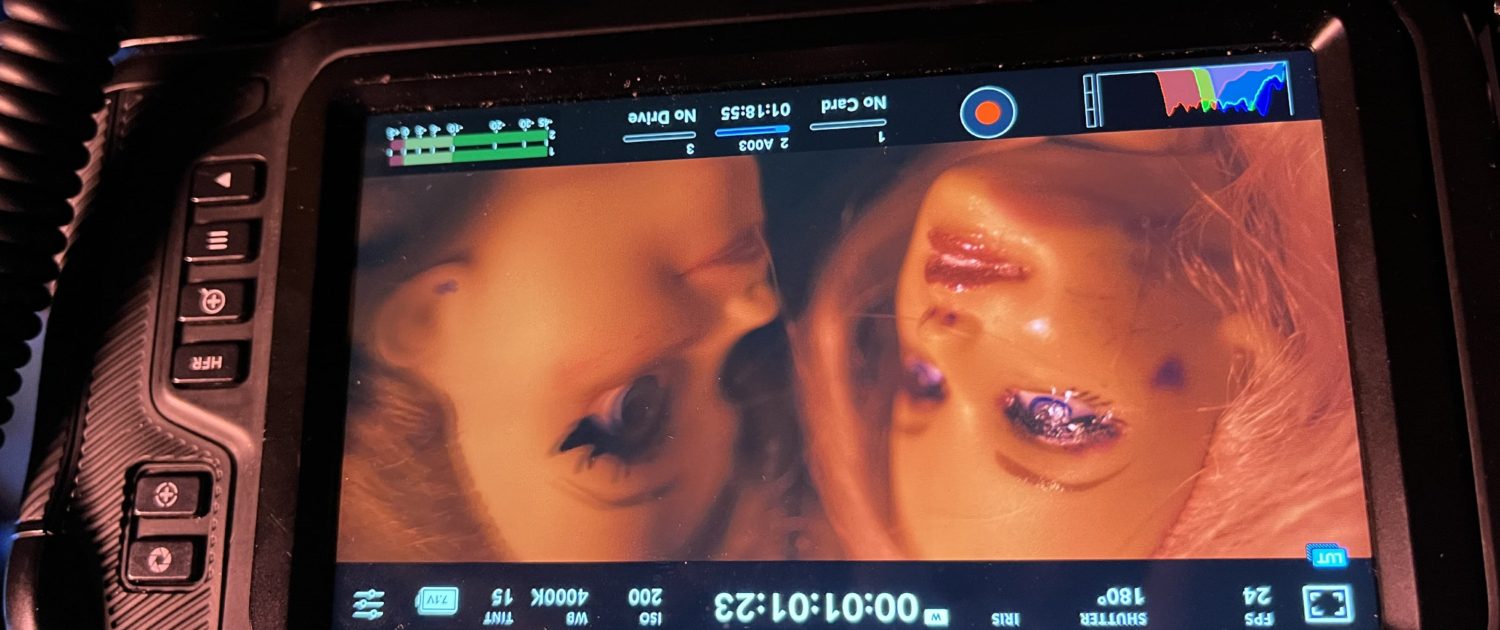 The image shows a camera monitor displaying two close-up faces of persons with dramatic makeup, set against a dark background with orange lighting.