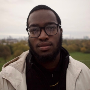 A person with glasses and a beard faces the camera, blurred city skyline and greenery in the background, overcast sky, natural daylight setting.