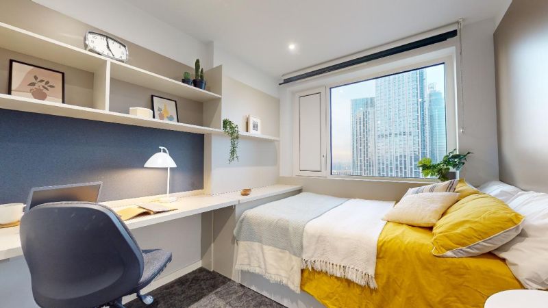 Modern bedroom with a white and yellow color scheme, study area with chair, shelves with decor, large window with city view, and green plants.