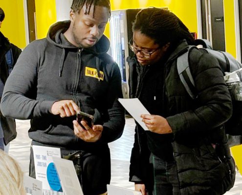 Two people are engaged in a conversation at a booth with flyers, one holding a smartphone and the other reading a pamphlet, possibly at a job fair.