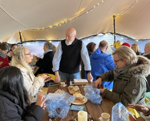 People gather under a lit tent, engaging in conversation, and food sharing. The atmosphere seems casual and convivial, with a focus on interaction and communion.
