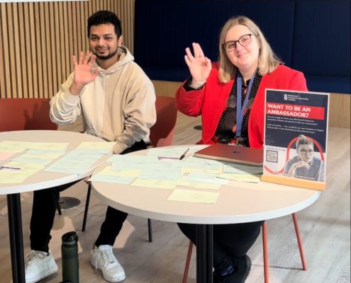 Two people are sitting at a table with papers, smiling and making peace signs, next to a sign about becoming an ambassador.