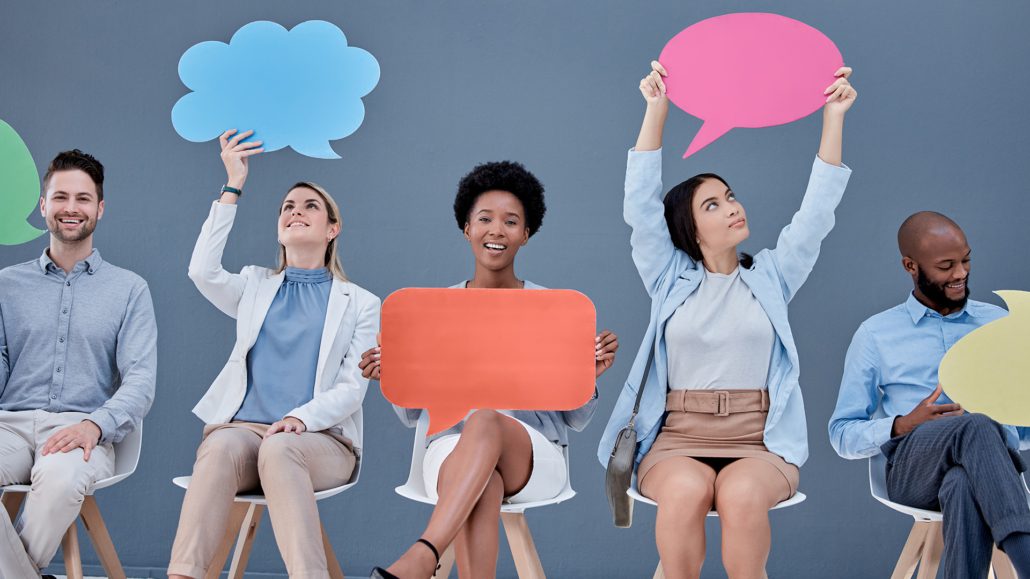 Five people sitting in a row, holding colorful speech bubble cutouts against a grey background, possibly portraying a concept of communication or diversity.