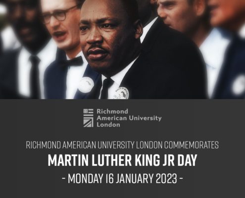 A smiling gentleman wearing a suit and bow tie stands proudly in front of a poster commemorating Martin Luther King Jr. Day at Richmond American University London.