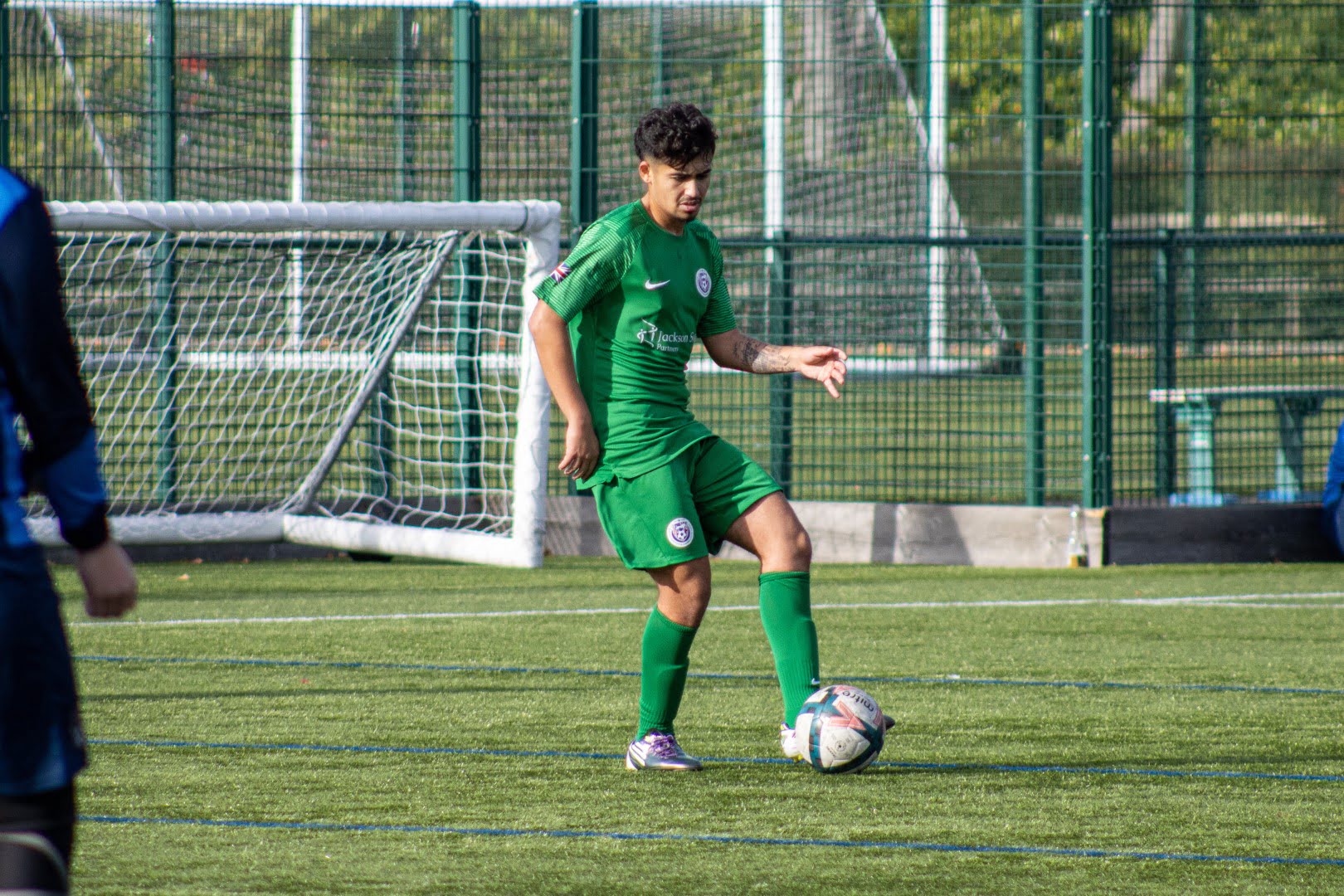 A soccer player in green attire controls the ball on an artificial pitch with a goal and fence in the background and another player partially visible.