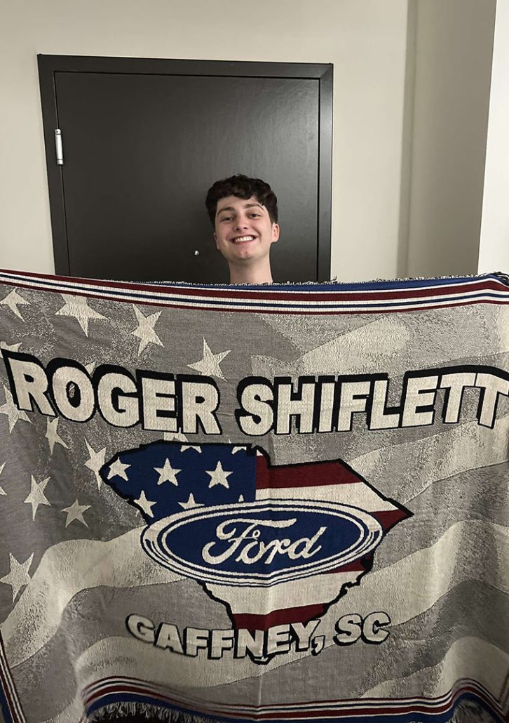 A smiling person is holding up a large blanket with a patriotic design featuring the "Ford" logo, stars, and text "Roger Shiflett Gaffney, SC".