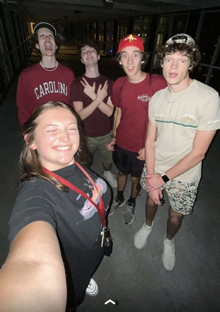 Five young adults pose for a selfie at night, with one person at the front smiling with eyes closed, and four others in the background making playful faces.