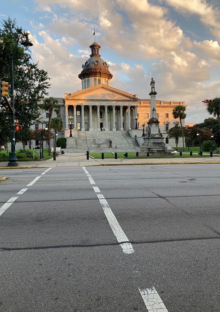 A domed building, possibly a capitol, with steps and statues at sunset. A pedestrian crossing and streetlamps frame the scene. Few people are visible.