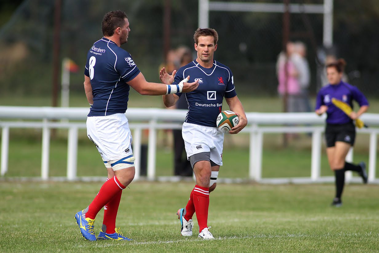 Two rugby players in blue jerseys are on a field; one with a rugby ball prepares to pass to the other. A referee in purple runs in the background.
