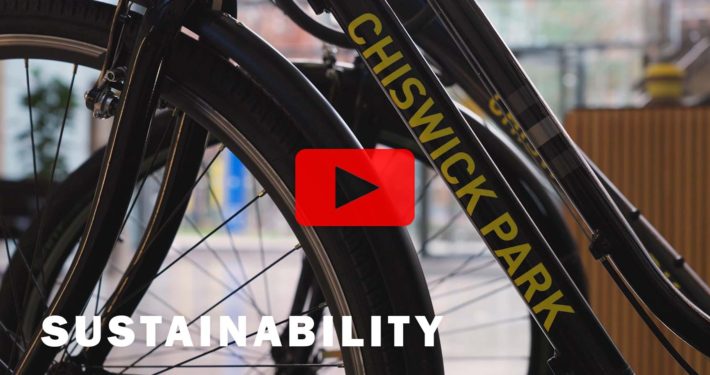 A cyclist is riding a bicycle with a carbon frame, a wheel rim, a tire, a spoke, a hub, a chain, a derailleur gear, a crankset, and other equipment, navigating the roads of Chiswick Park Sustainability.
