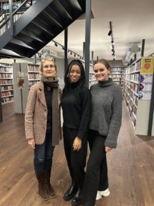 Three people pose smiling in a library, with shelves of books, a staircase, and modern lighting fixtures in the background. They appear friendly and casual.