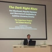 In this image, Dr. Caleb Turner is presenting on the topic of Dark Knight Rises, Populist Heroism, and Übermensch Aesthetics in Sublime-Uncanny Cinema at the 3rd Global Meeting of The Superhero Project. Full Text: The Dark Right Rises Bat-Manhood, Populist Heroism and Übermensch Aesthetics in Sublime-Uncanny Cinema The Superhero Project 3rd Global Meeting Dr. Caleb Turner Assistant Professor in Film Studies Richmond University, The American University in London