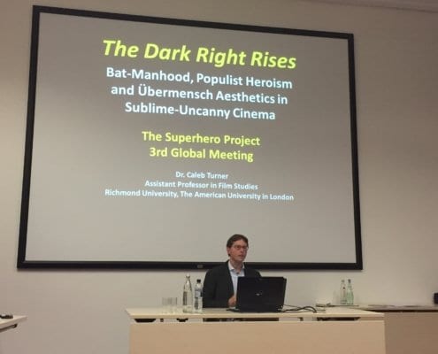 In this image, Dr. Caleb Turner is presenting on the topic of Dark Knight Rises, Populist Heroism, and Übermensch Aesthetics in Sublime-Uncanny Cinema at the 3rd Global Meeting of The Superhero Project. Full Text: The Dark Right Rises Bat-Manhood, Populist Heroism and Übermensch Aesthetics in Sublime-Uncanny Cinema The Superhero Project 3rd Global Meeting Dr. Caleb Turner Assistant Professor in Film Studies Richmond University, The American University in London