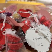 The image shows a collection of red heart-shaped chocolates with personalized notes attached, expressing love and affection, possibly for Valentine's Day.